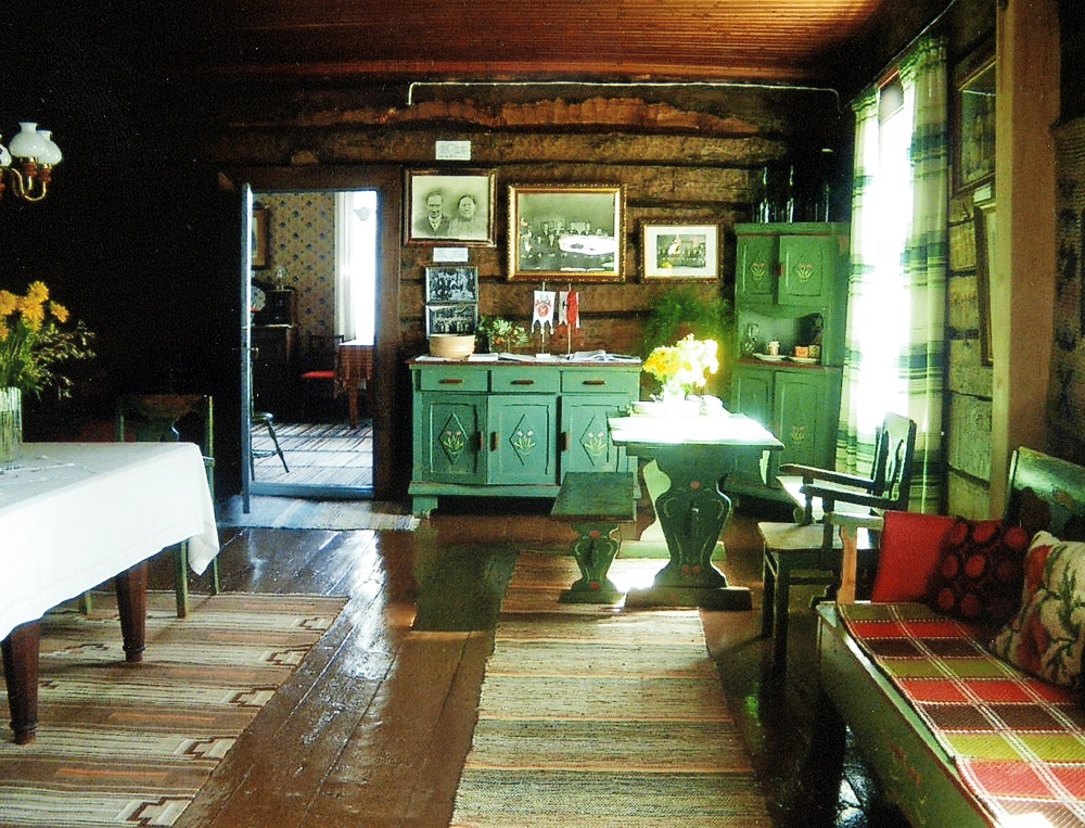 Tuvan sisustus. Ruskeaksi maalatut lattialankut, paljaat hirsiseinät, Vihreäksi maalattuja kalusteita: senkki, kaappi, puusohva ja pöytä penkkeineen. Taustalla avoin ovi ja näkymä toiseen huoneeseen.