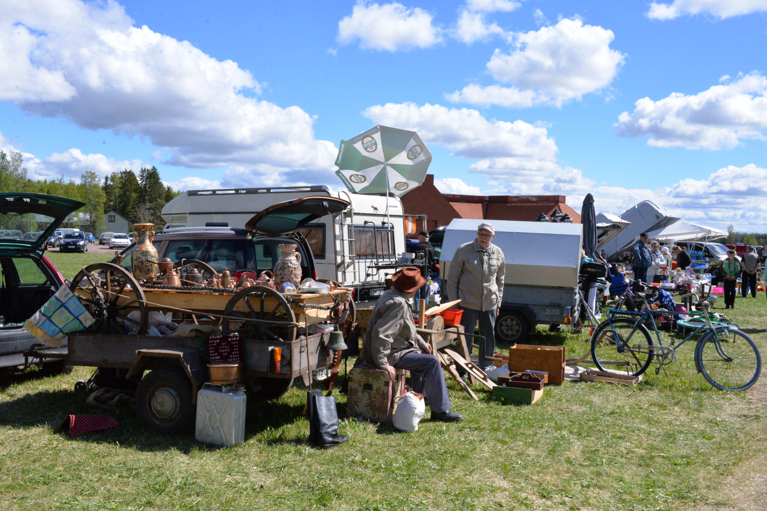 Kaksi miestä juttelevat, taustalla autoja ja takakontteja täynnä erilaista tavaraa jota he myyvät. Polkupyöriä, kärrynpyöriä, astioita, kirjoja. Nurmikko vihertää ja aurinko paistaa.